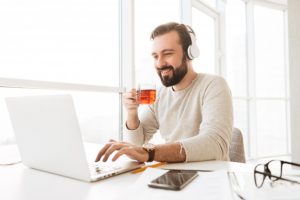 european-joyous-man-with-short-brown-hair-drinking-tea-listening-music-via-wireless-headphones-while-using-notebook_171337-27088